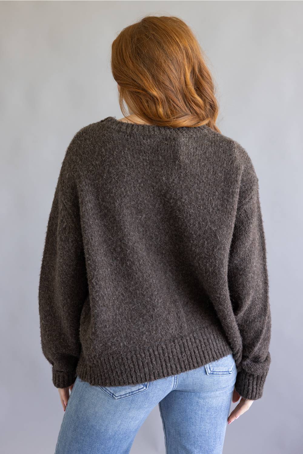 A woman with red hair is shown from behind wearing a dark brown fuzzy knit sweater and light blue jeans against a plain gray background.