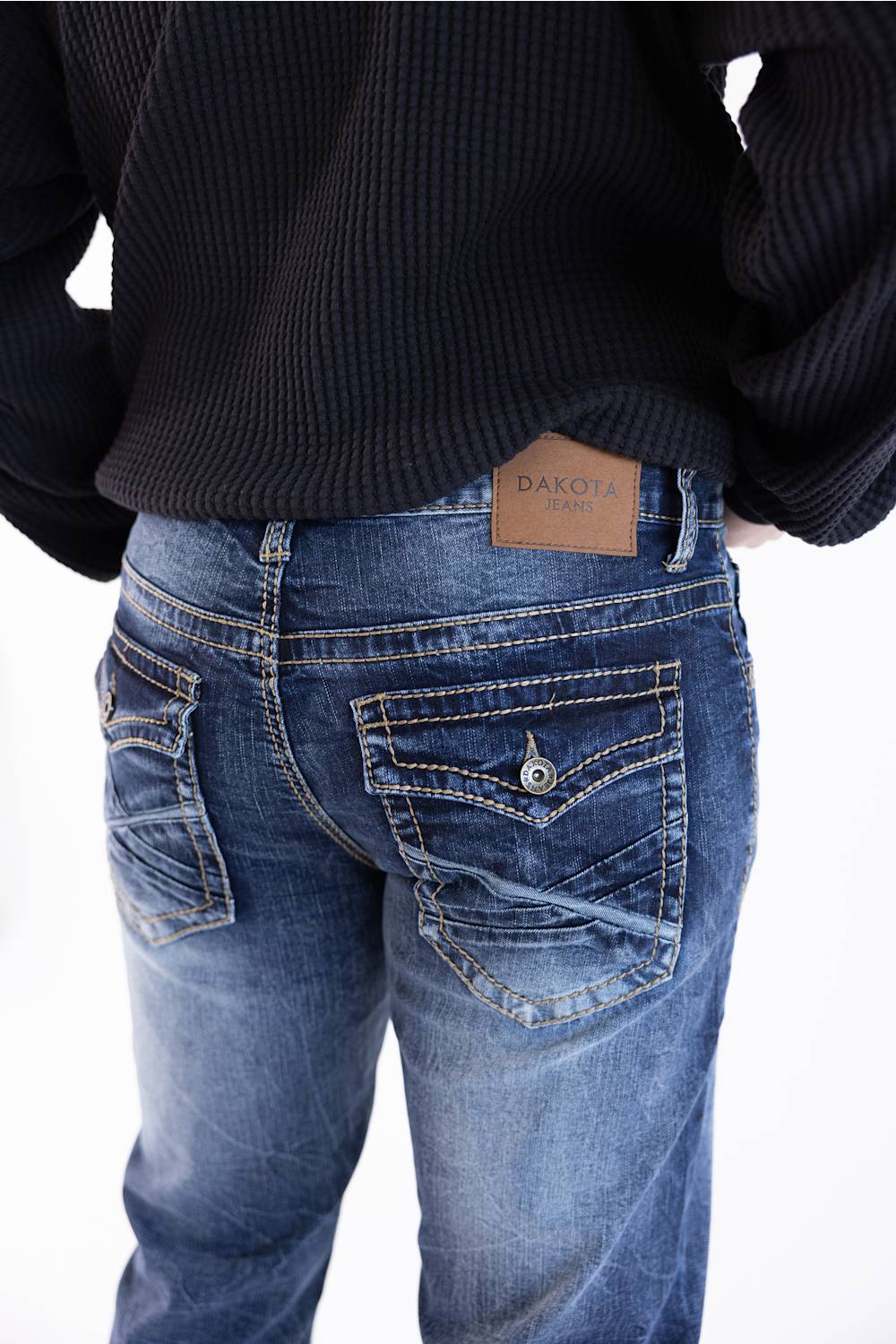 Rear view of a person wearing blue denim jeans with a tan leather DAKOTA brand patch at the back waistband, paired with a black ribbed sweater against a white background.