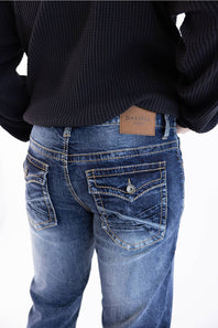 Rear view of a person wearing blue denim jeans with a tan leather DAKOTA brand patch at the back waistband, paired with a black ribbed sweater against a white background.