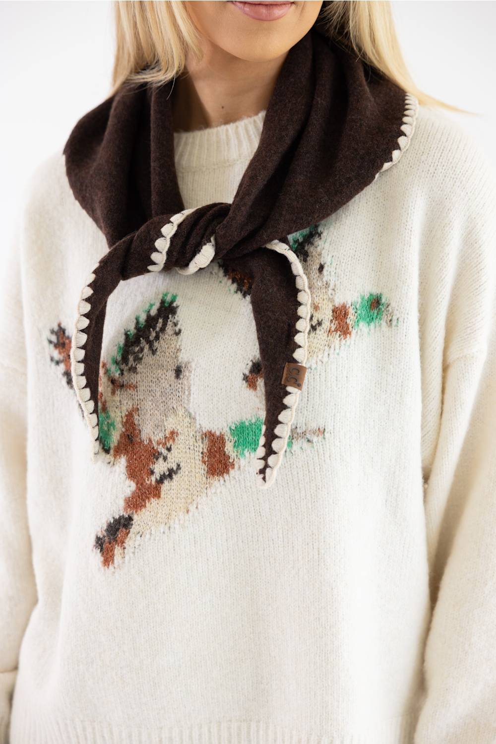A close-up of a person wearing a cream knit sweater with a large embroidered bird motif in brown, green, and beige, paired with a dark brown scarf tied around the neck.