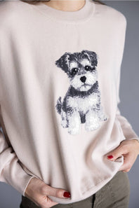 A close-up of a person wearing a light blush pink crewneck sweater with a sequined dog graphic on the front, paired with olive green pants.