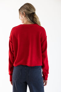 A woman viewed from the back wearing a red sweater and dark blue jeans with pinstripes standing in front of a white studio background