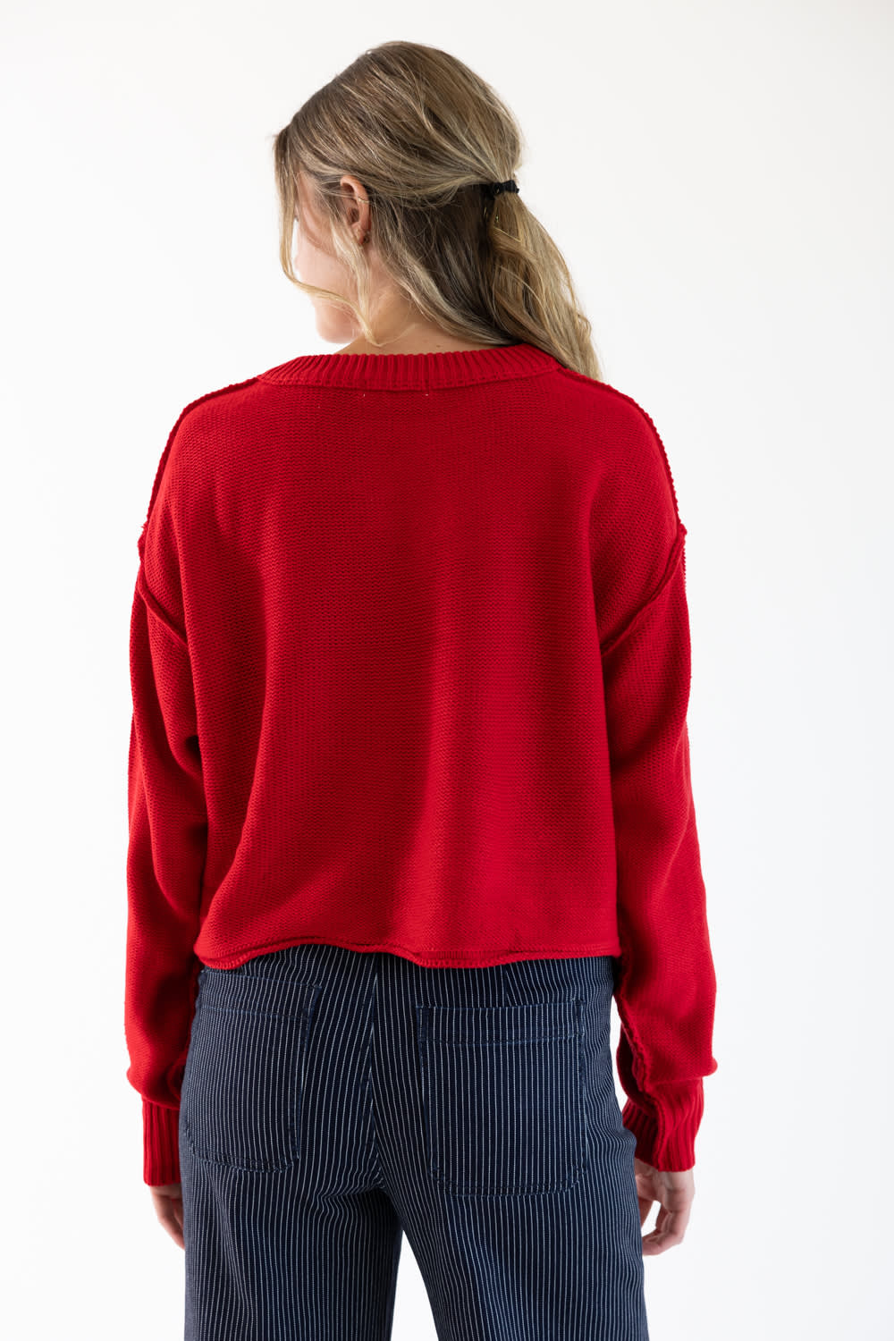 A woman viewed from the back wearing a red sweater and dark blue jeans with pinstripes standing in front of a white studio background