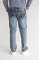 Rear view of a person wearing light-wash blue jeans with red and black contrast stitching on the back pockets, paired with a gray ribbed sweater and white sneakers, against a plain light gray studio background.