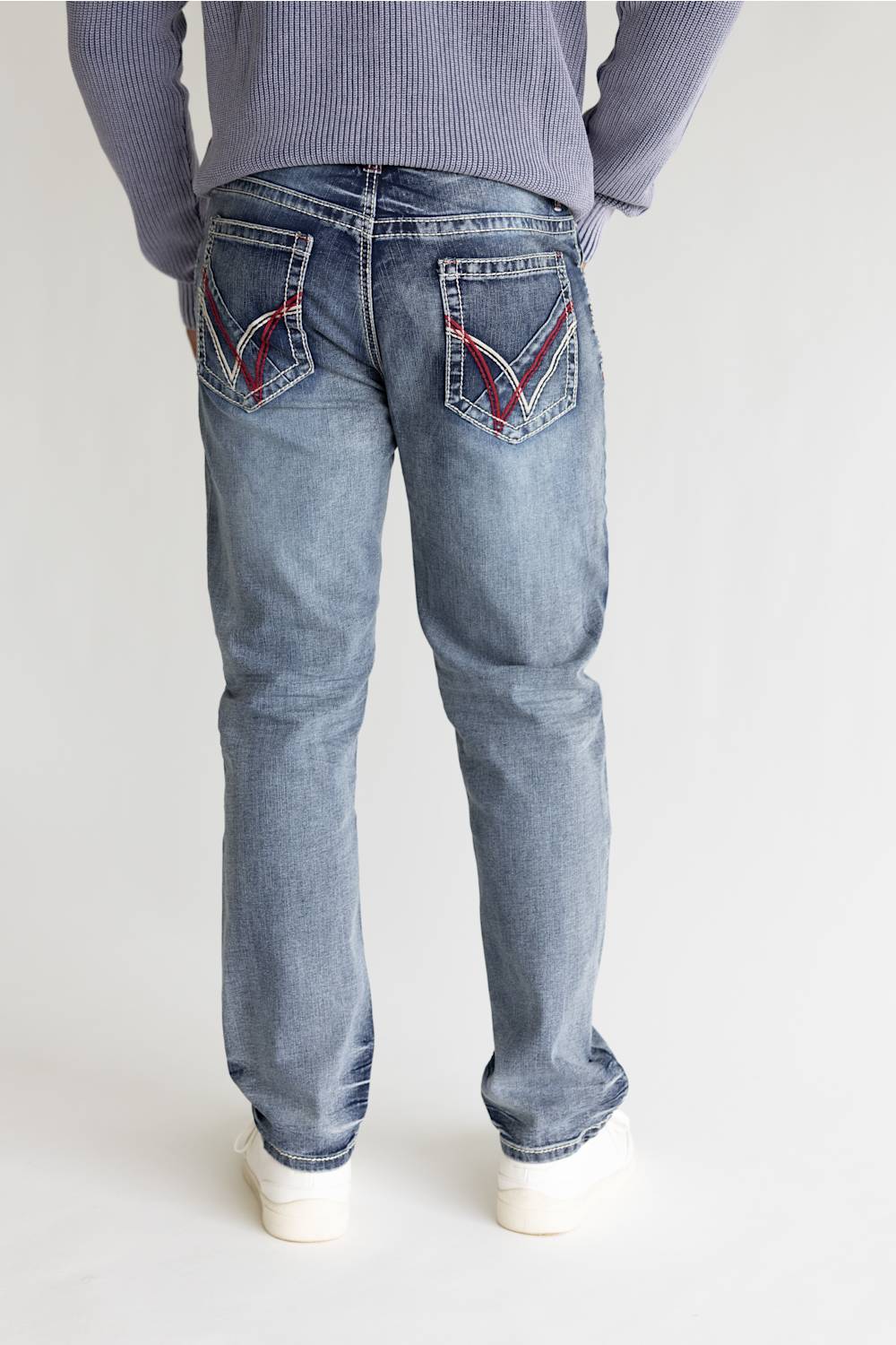 Rear view of a person wearing light-wash blue jeans with red and black contrast stitching on the back pockets, paired with a gray ribbed sweater and white sneakers, against a plain light gray studio background.