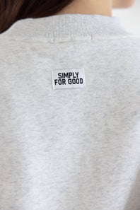 Close-up of a person wearing a light heather gray crewneck sweatshirt, showing a white rectangular brand patch on the upper back with the text 'SIMPLY FOR GOOD'.