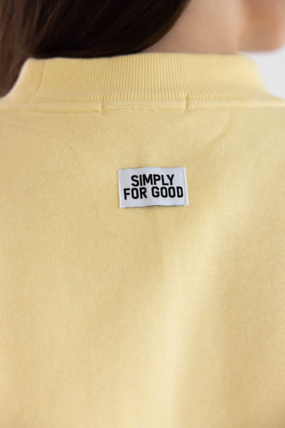 Close-up of the back of a pale yellow sweatshirt with a white rectangular brand patch reading 'SIMPLY FOR GOOD' on the upper back.
