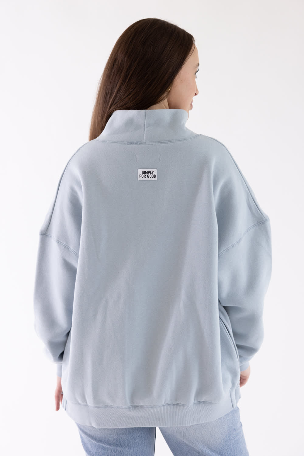 A woman shown from the back wearing a light blue sweatshirt with a small white rectangular logo patch on the upper back, paired with light blue jeans against a plain white studio background.
