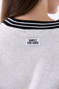 Close-up back view of a person wearing a light heather gray crewneck sweatshirt with black and white striped ribbed collar trim and a white rectangular patch reading 'SIMPLY FOR GOOD' on the upper back.