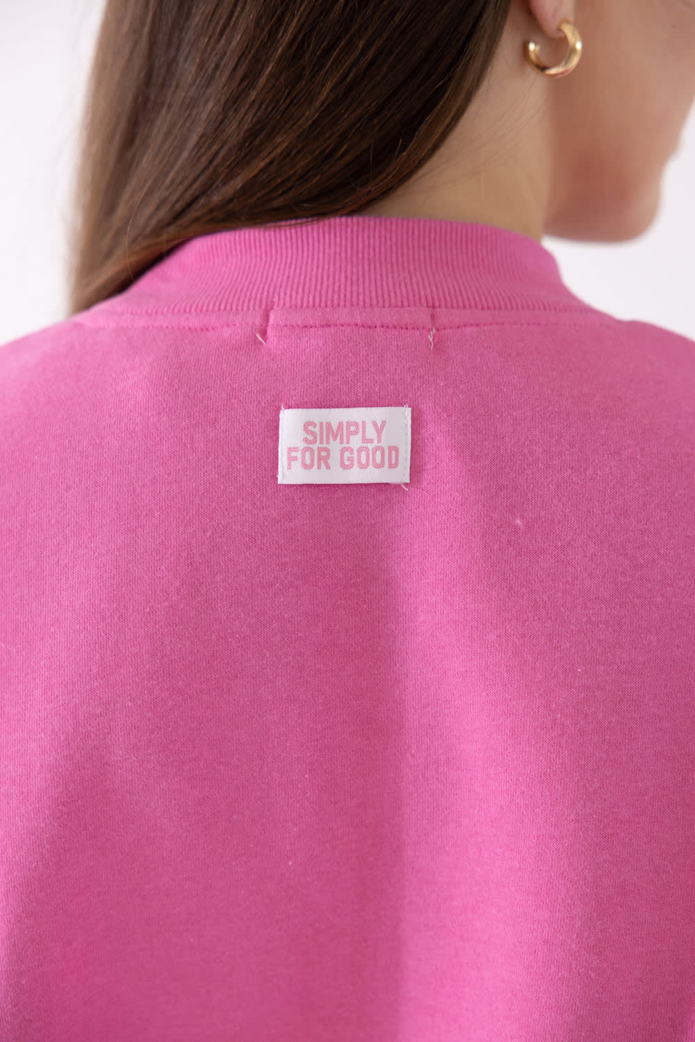 Close-up back view of a person wearing a bright pink crewneck sweatshirt with a small white rectangular patch reading 'SIMPLY FOR GOOD' on the upper back.