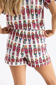 A woman wearing a white romper with an all-over colorful nutcracker soldier print, shown from the shoulders to mid-thigh against a plain white studio background.