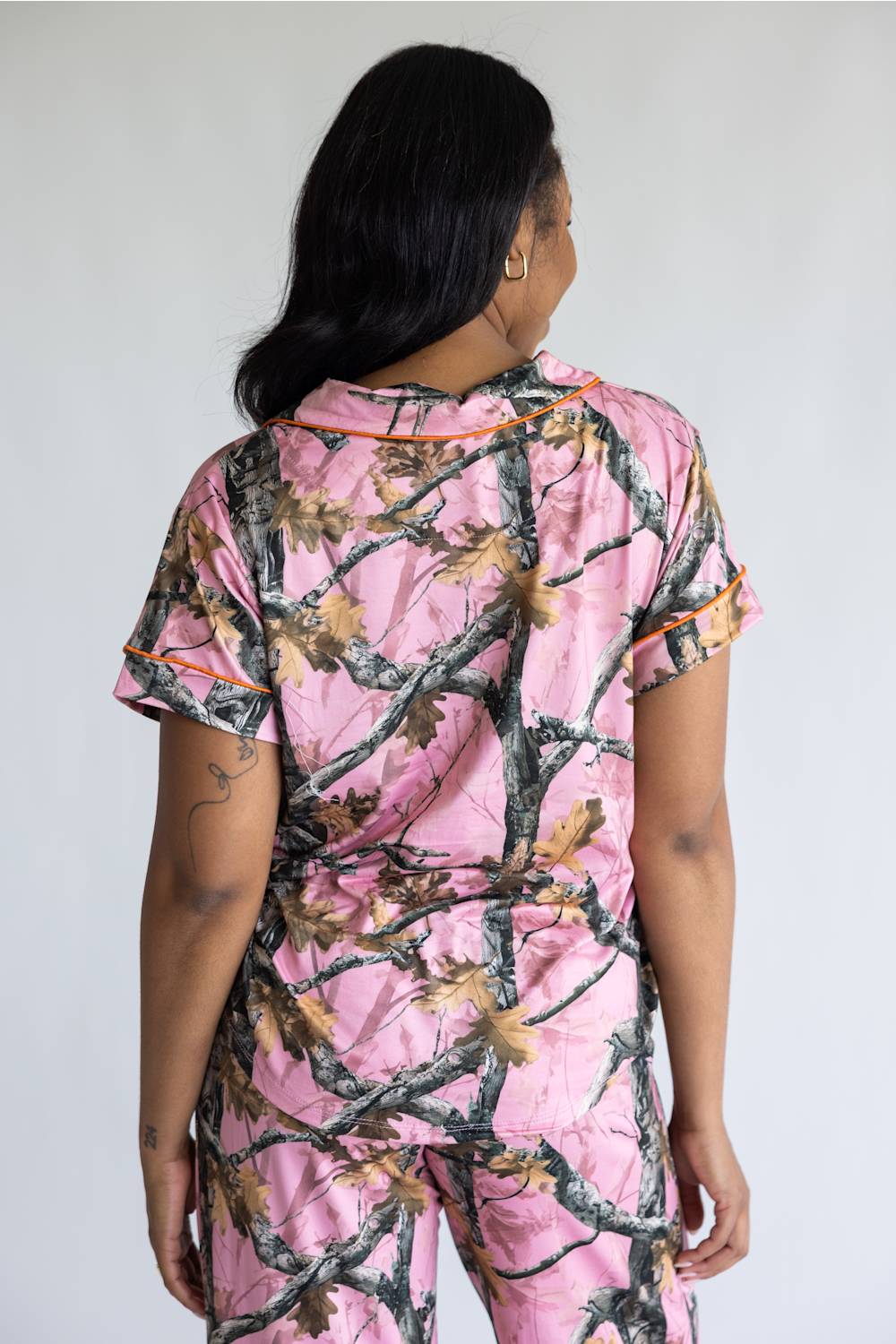 A woman viewed from behind wearing a pink short-sleeve pajama set with a woodland camouflage branch and leaf print, featuring thin orange piping along the collar and sleeve cuffs.