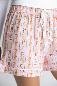 Pink drawstring shorts with a vertical striped pattern featuring tan bows and small dog illustrations, shown on a model.