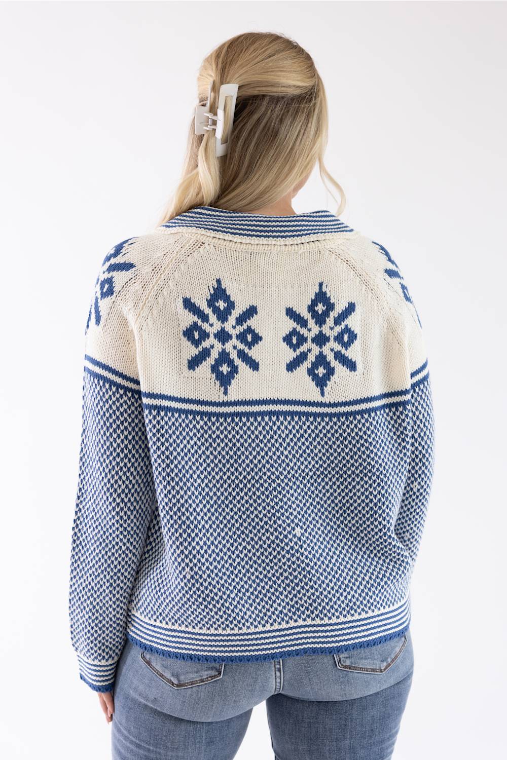 A woman viewed from the back wearing a blue and cream knit cardigan with geometric snowflake patterns, paired with blue jeans against a plain white background.
