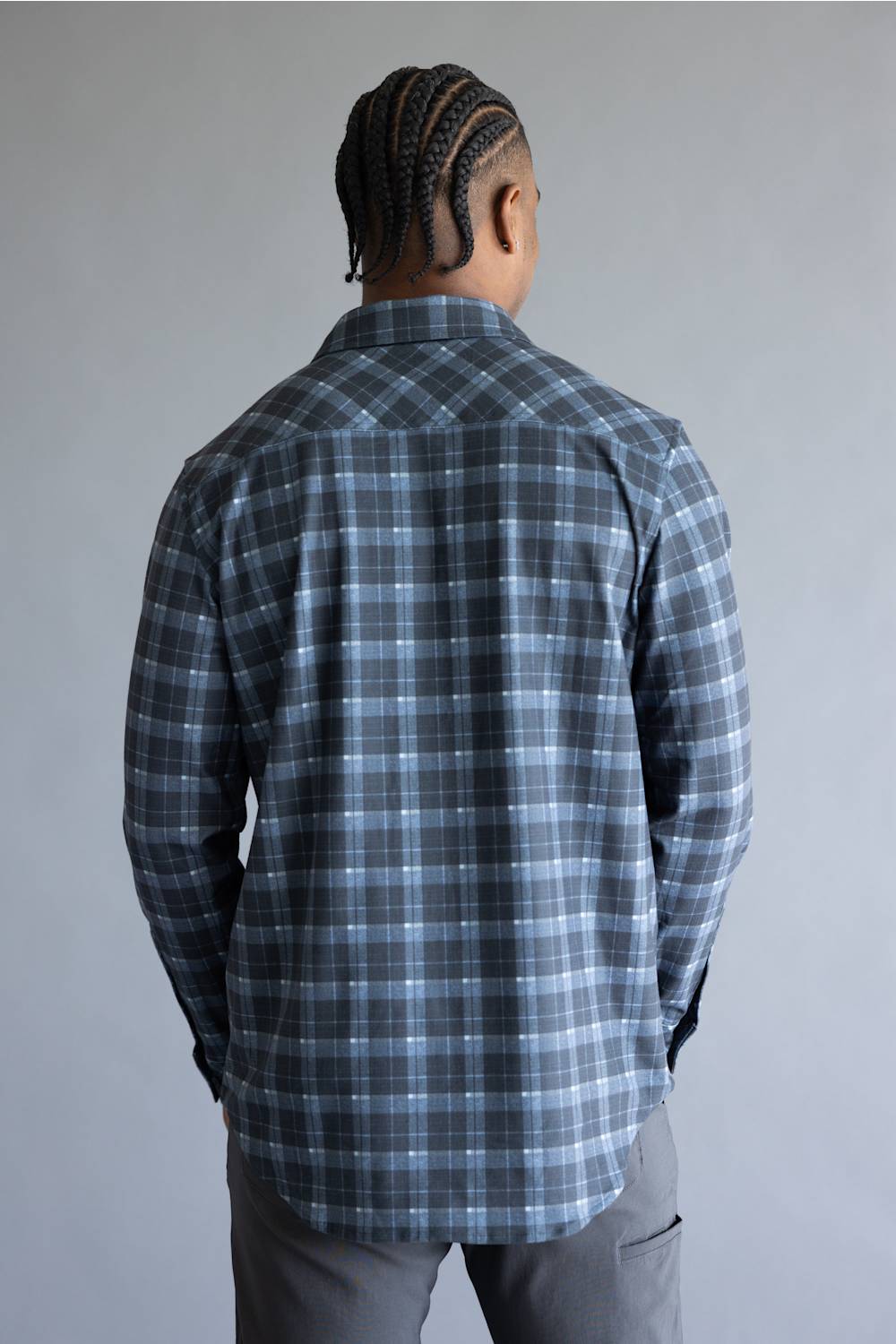 A man viewed from behind wearing a long-sleeve plaid button-up shirt in dark gray and light blue, paired with gray pants, against a plain gray studio background.