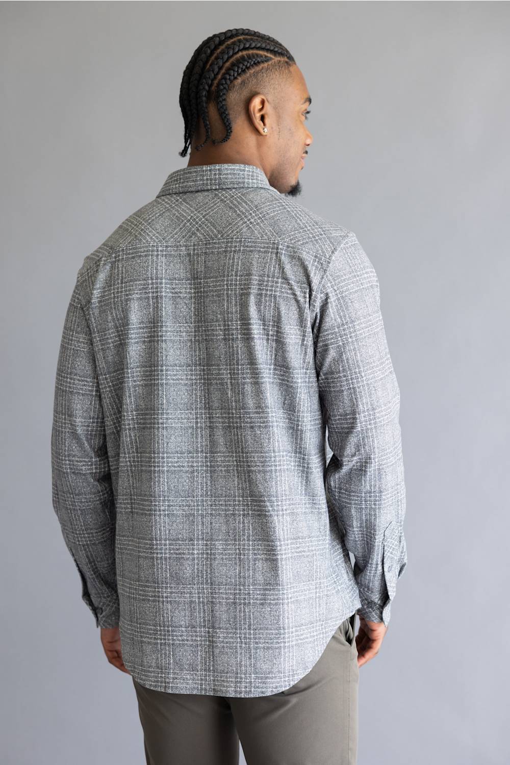 A man viewed from the back wearing a light gray and white plaid button-down shirt and gray pants, standing against a solid gray studio background.