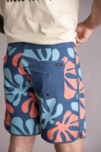 Back view of a person wearing blue shorts with colorful leaf pattern on a plain background. The shorts are named Quiksilver Sol Tiles 19" Boardshorts for Men in Dark Denim.