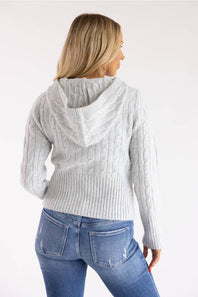 A woman shown from the back wearing a light gray cable-knit hooded sweater and blue jeans against a plain white studio background.