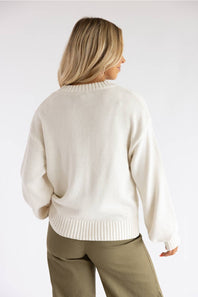 A woman shown from the back wearing a cream-colored knit sweater and olive green pants against a plain white studio background.