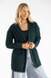 A woman wearing a dark forest green cable-knit cardigan over a black top and light blue jeans, photographed against a clean white studio background.
