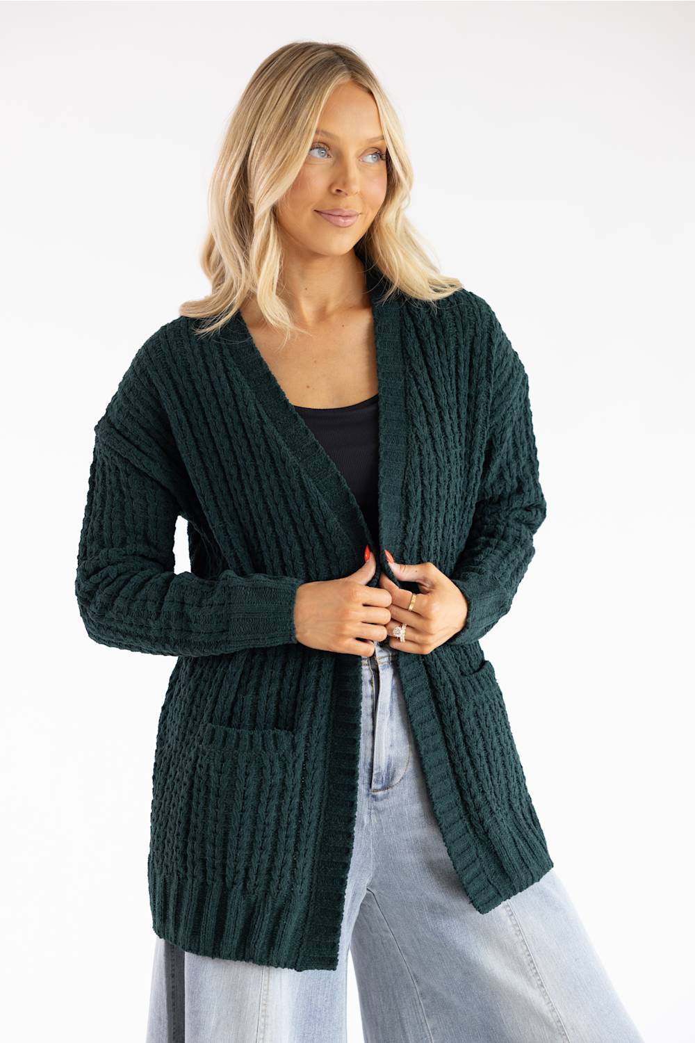 A woman wearing a dark forest green cable-knit cardigan over a black top and light blue jeans, photographed against a clean white studio background.