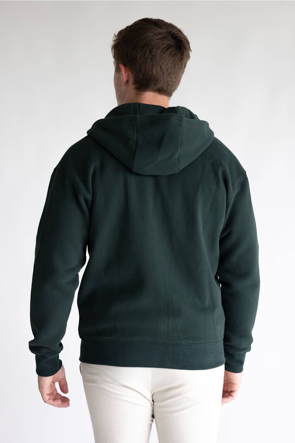 A man viewed from behind wearing a dark forest green pullover hoodie and white pants, standing against a plain light gray studio background.