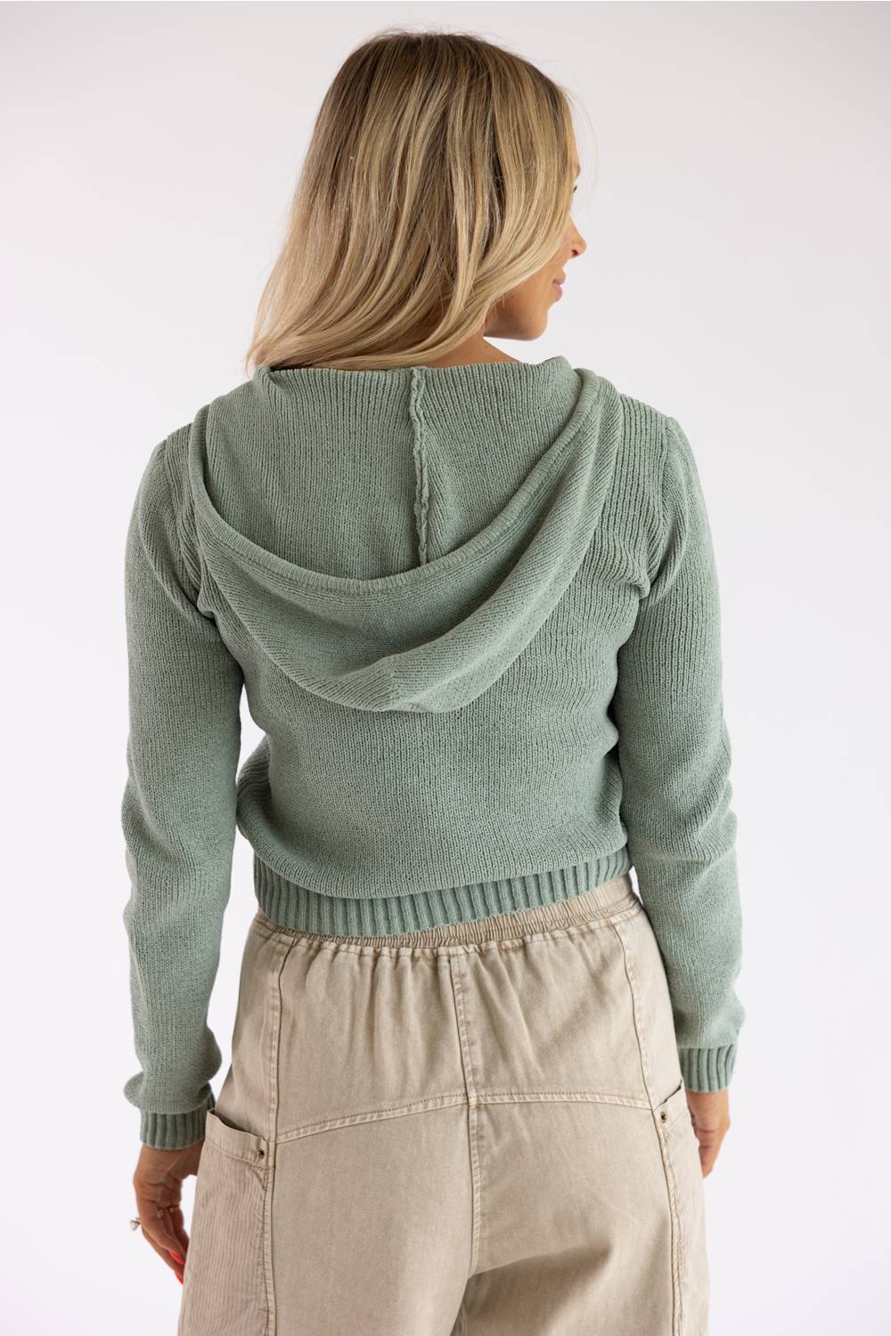 A woman shown from the back wearing a sage green ribbed knit hooded sweater and beige pants against a plain light gray studio background.