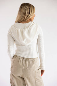 Back view of a model wearing a Pink Rose blizzard white cable-knit full-zip sweater hoodie styled with beige drawstring utility cargo pants against a plain backdrop.