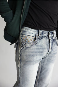 Close-up of a person wearing light-wash blue denim jeans with a decorative gold geometric pattern on the back pocket, styled with a black t-shirt and a dark green zip-up hoodie.