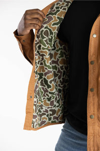 A close-up of a person wearing a tan overshirt with a green, brown, and beige camouflage pattern lining, layered over a black top and blue jeans.