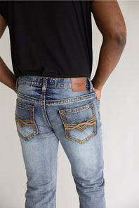 Rear view of a person wearing light-wash blue jeans with decorative gold contrast stitching and a brown leather patch on the back waistband, paired with a black shirt against a plain light background.