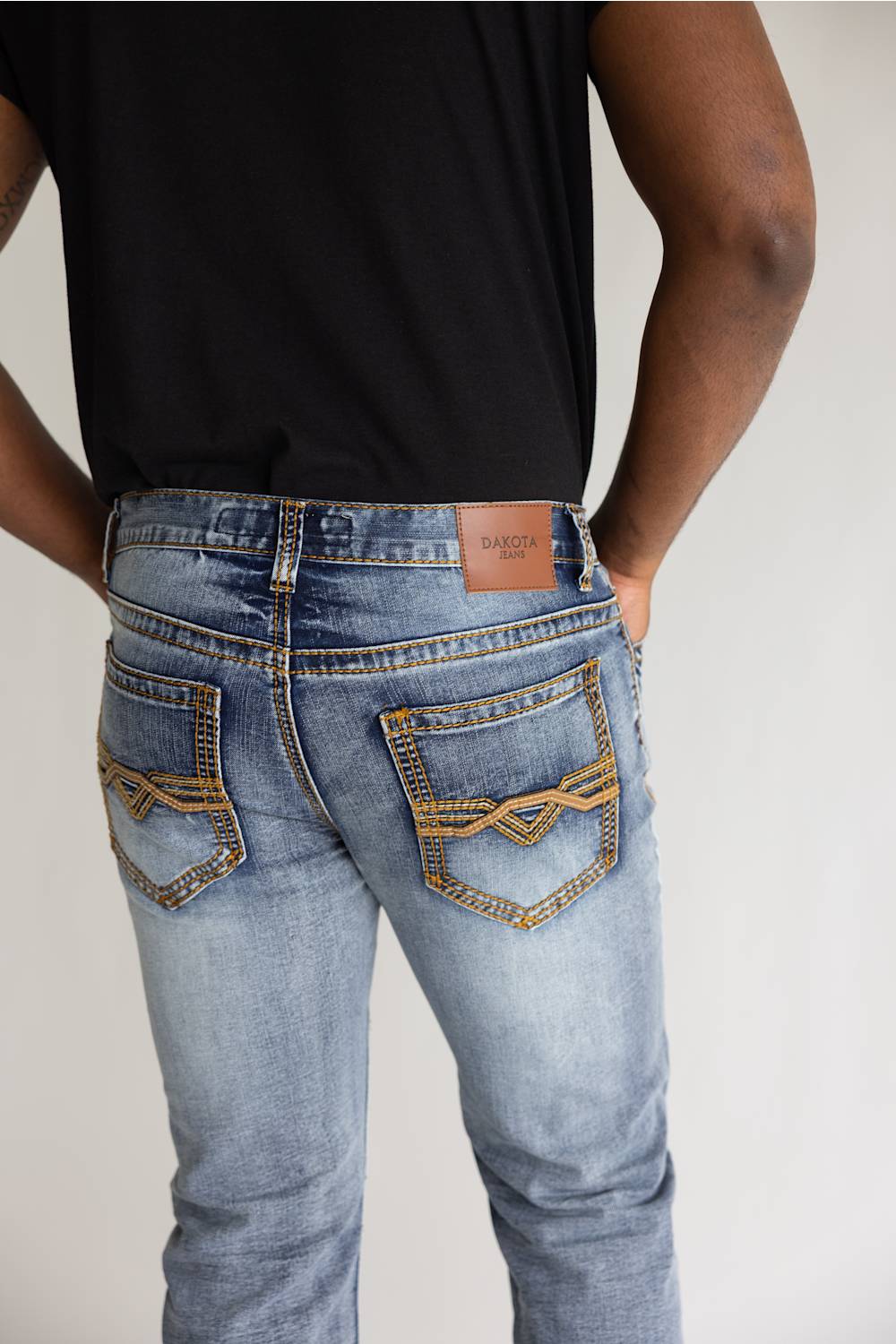 Rear view of a person wearing light-wash blue jeans with decorative gold contrast stitching and a brown leather patch on the back waistband, paired with a black shirt against a plain light background.