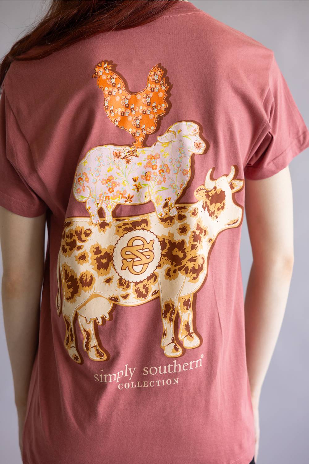 A person wearing a dusty rose short-sleeve T-shirt, shown from the back. The shirt features a large graphic print of a cow, sheep, and chicken in cream, orange, and brown tones, with the text 'simply southern collection' printed below.