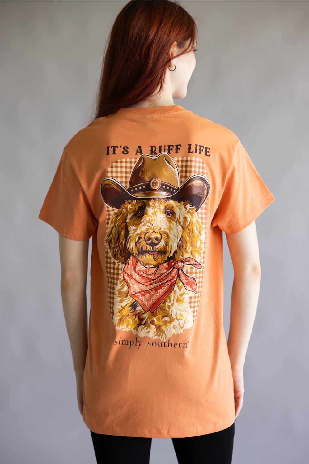 A woman shown from the back wearing a peach-colored graphic t-shirt. The shirt features a large print of a curly-haired dog wearing a cowboy hat and bandana, with the text 'IT'S A RUFF LIFE' and 'simply southern!' printed around the graphic.