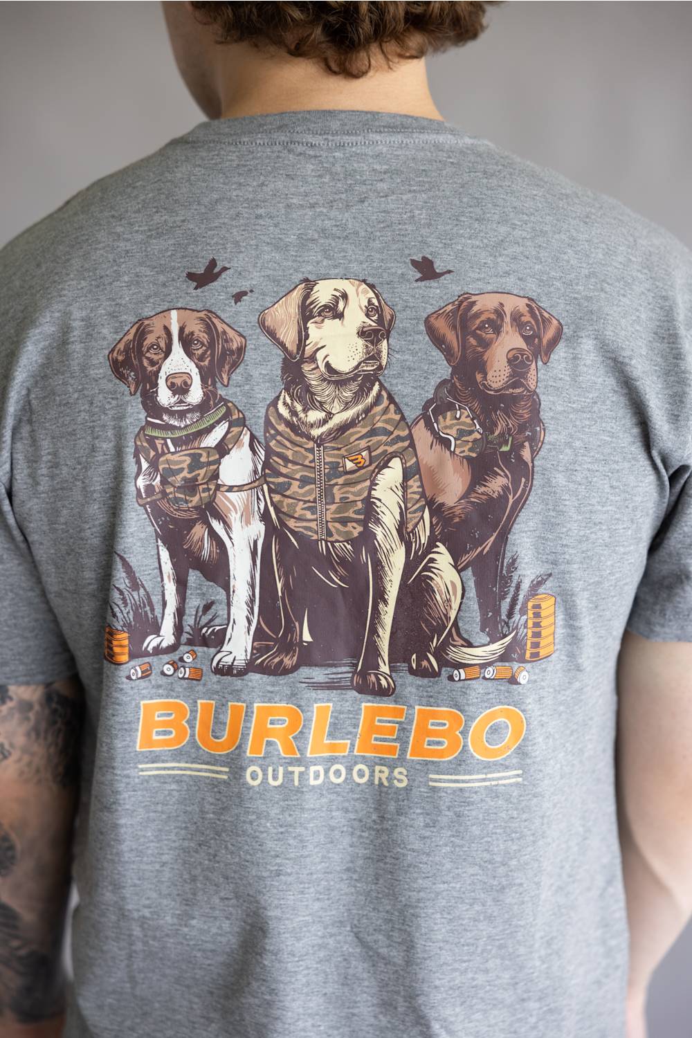 A person viewed from the back wearing a heather gray t-shirt with a large graphic print of three dogs in camouflage hunting vests and the text 'BURLEBO OUTDOORS' in orange and white.