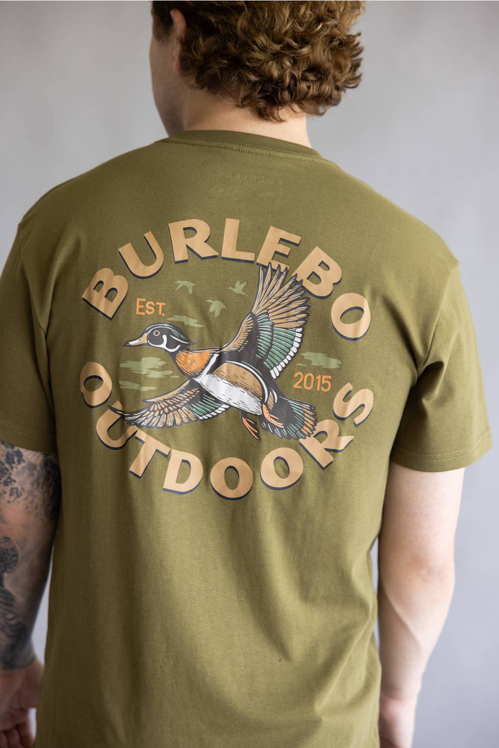 A man viewed from behind wearing an olive green t-shirt with a large graphic on the back. The graphic features the text 'BURLEBO OUTDOORS' in a tan circular font surrounding an illustration of a flying mallard duck with 'Est.