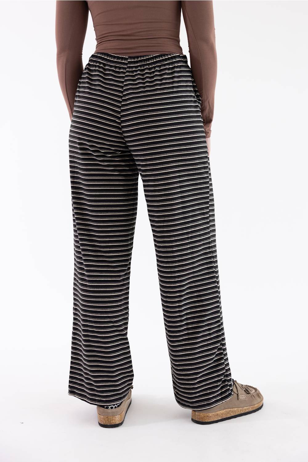 A person shown from the back wearing black and white horizontally striped wide-leg pants, a brown top, and tan slip-on shoes against a plain white studio background.