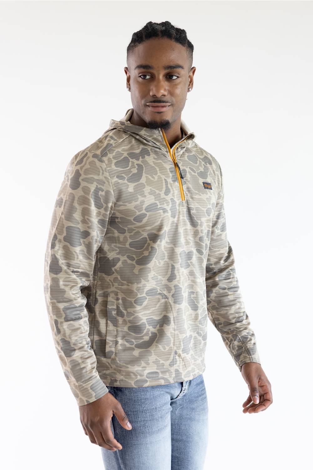 A man wearing a beige and gray camouflage quarter-zip hoodie with a yellow zipper pull, paired with blue jeans, photographed against a plain white studio background.