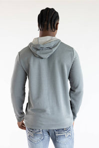 A man viewed from behind wearing a light gray ribbed hooded sweatshirt and light blue jeans with decorative stitching on the back pockets, against a plain white studio background.