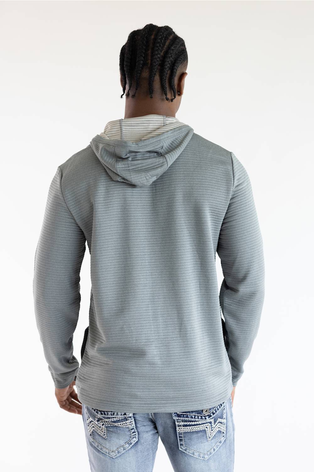 A man viewed from behind wearing a light gray ribbed hooded sweatshirt and light blue jeans with decorative stitching on the back pockets, against a plain white studio background.