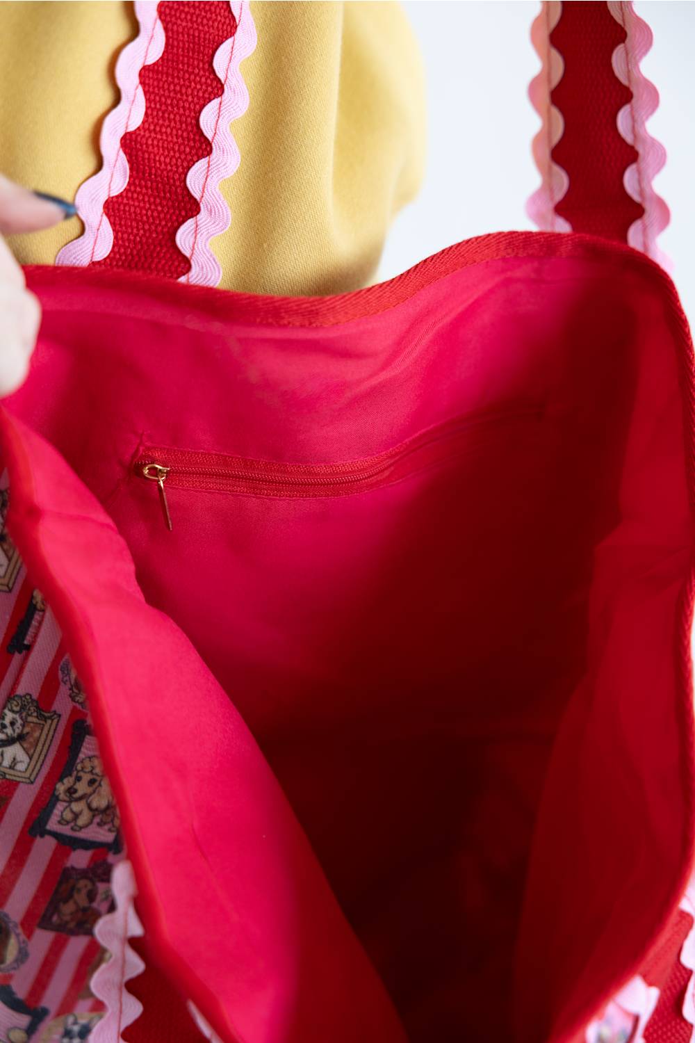 Close-up of a bright red fabric tote bag interior with a zippered pocket, pink scalloped trim, and part of a patterned exterior strap visible.