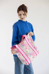 A woman wearing a blue sweatshirt and jeans holds a large woven tote bag with pink trim and a pink, white, and blue floral pattern against a plain white background.