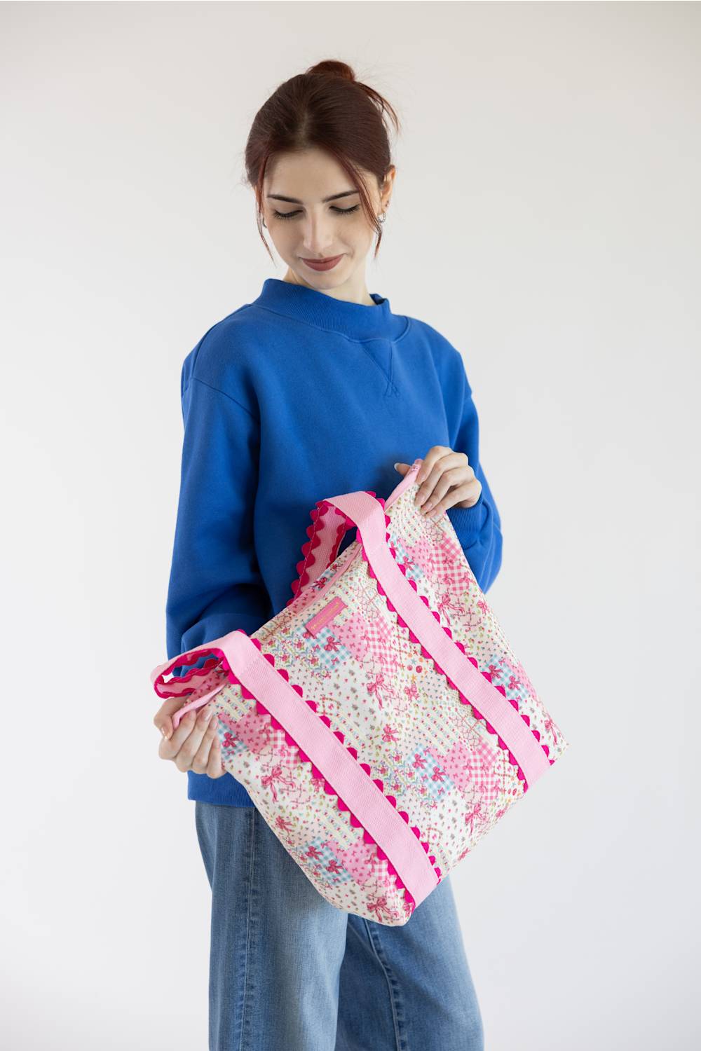 A woman wearing a blue sweatshirt and jeans holds a large woven tote bag with pink trim and a pink, white, and blue floral pattern against a plain white background.