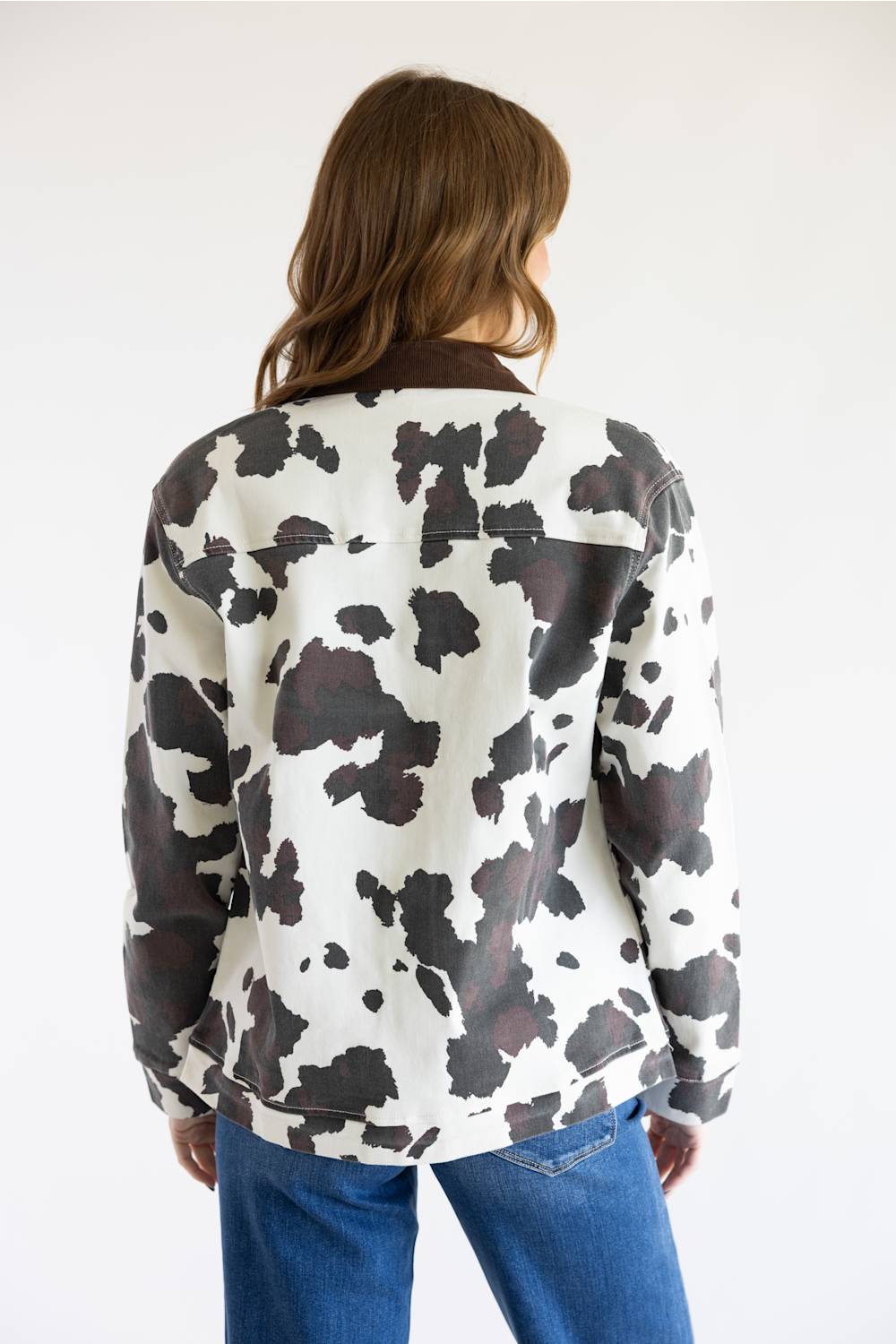 A woman viewed from behind wearing a cow-print jacket with dark brown collar and cuffs, paired with blue jeans, against a plain white background.