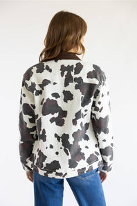 A woman viewed from behind wearing a cow-print jacket with dark brown collar and cuffs, paired with blue jeans, against a plain white background.