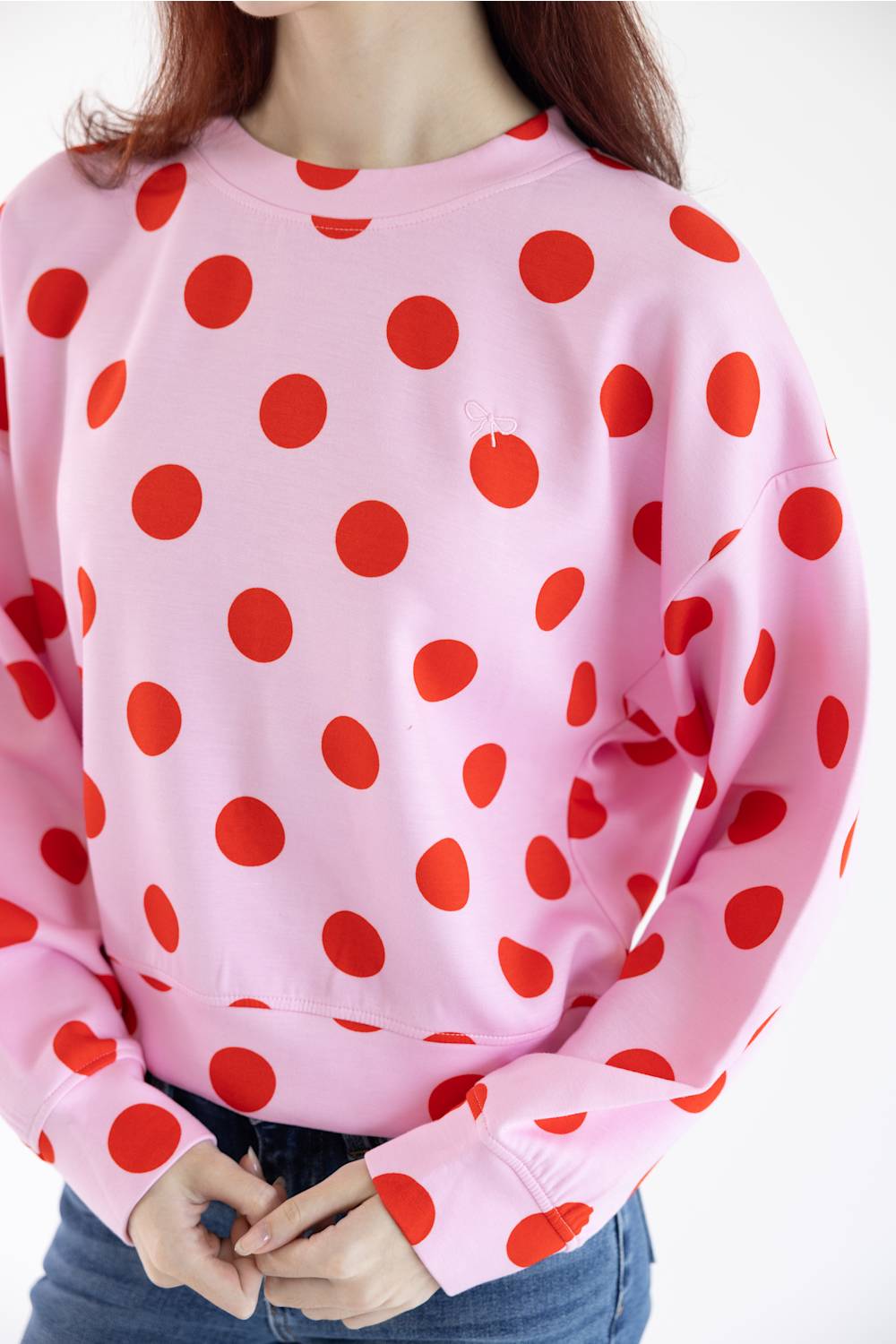 A person wearing a light pink crewneck sweatshirt with a bold red polka dot pattern, paired with blue jeans against a plain white background.