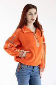 A woman modeling a bright orange sherpa fleece half-zip pullover sweater with patterned trim details on the collar, zipper placket, cuffs, and sleeves, paired with blue jeans against a plain white studio background.