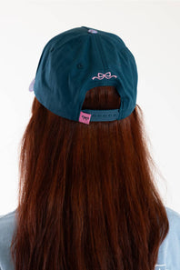 A person viewed from behind wearing a dark teal baseball cap with a pink bow graphic on the back and an adjustable strap. The person has long, straight reddish-brown hair and is wearing a light blue top against a plain white background.