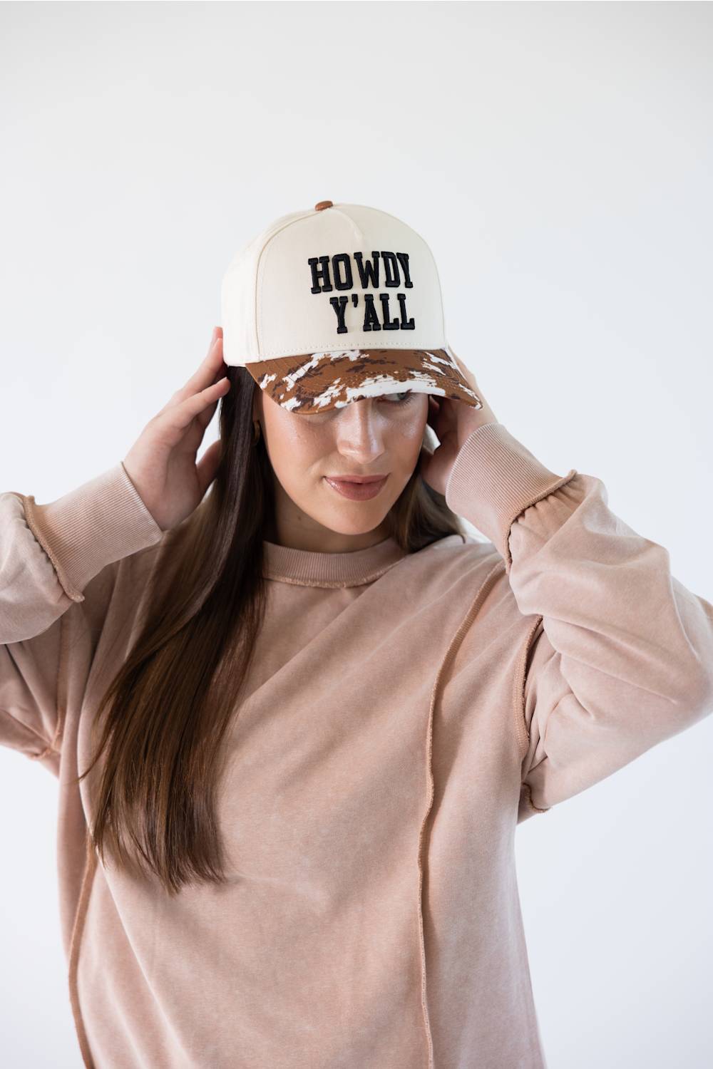 A woman wearing a beige trucker-style cap with 'HOWDY Y'ALL' embroidered on the front, paired with a matching beige sweatshirt, set against a plain light gray background.