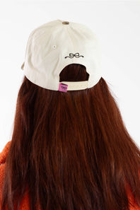 Back view of a person with long brown hair wearing an off-white baseball cap with a small black bow-tie graphic and a pink brand tag.